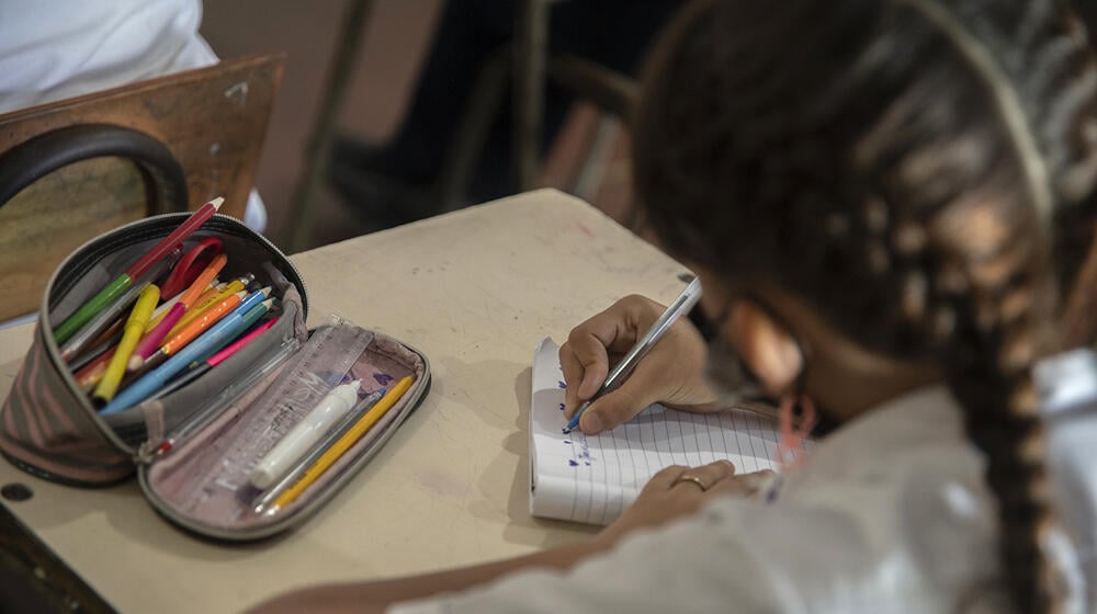 Niña estudiante escribiendo en pupitre en aula de escuela. 