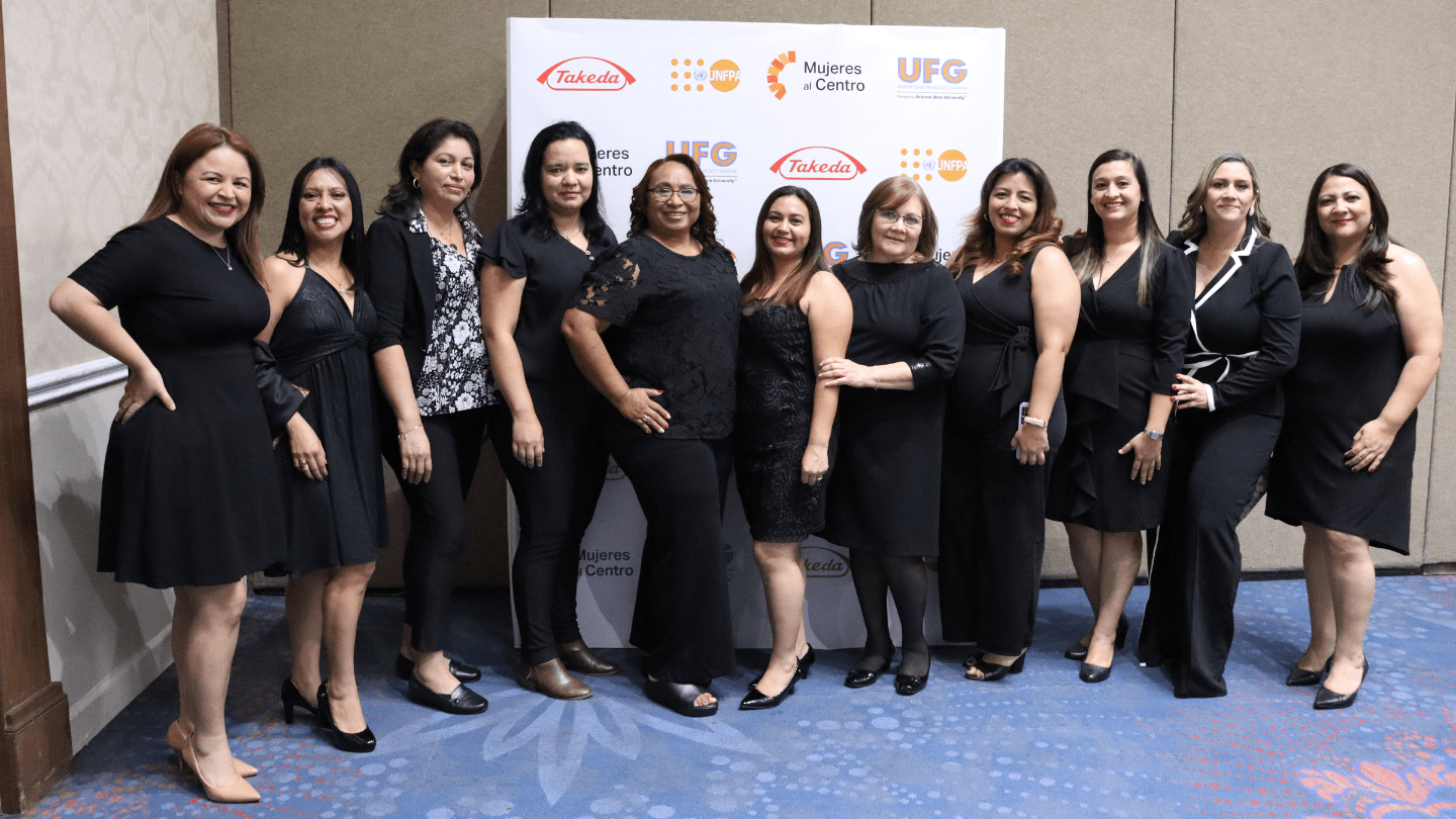 Once mujeres posan juntas, vistiendo de negro, frente a un fondo con los logotipos de Takeda, UNFPA, Mujeres al Centro y UFG. Se ven sonrientes y unidas, reflejando un ambiente de celebración y empoderamiento.