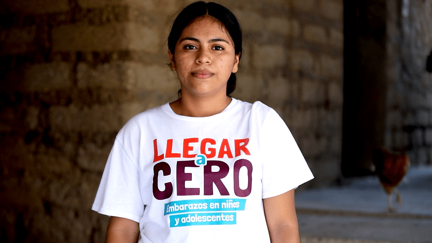Katherine, en el patio de su casa, viendo de frente a la cámara. Utiliza una camisa con un estampado que dice: Llegar a Cero embarazos en niñas y adolescentes.